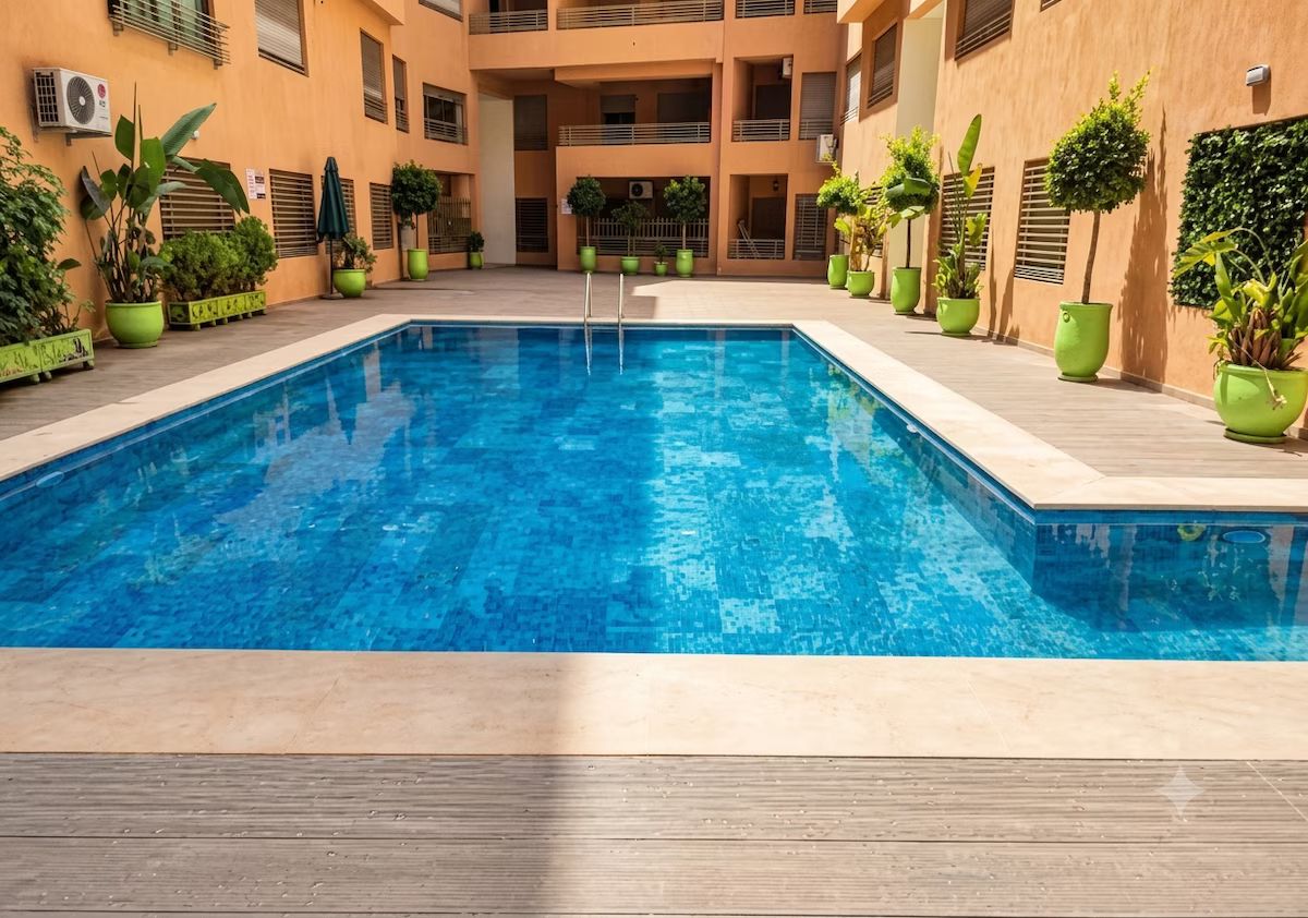 A swimming pool with a wooden deck, surrounded by potted plants and a multi-level building with balconies.