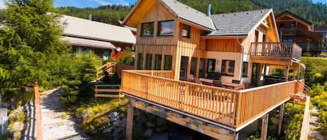 A wooden cabin with a balcony, surrounded by trees and a gravel path.