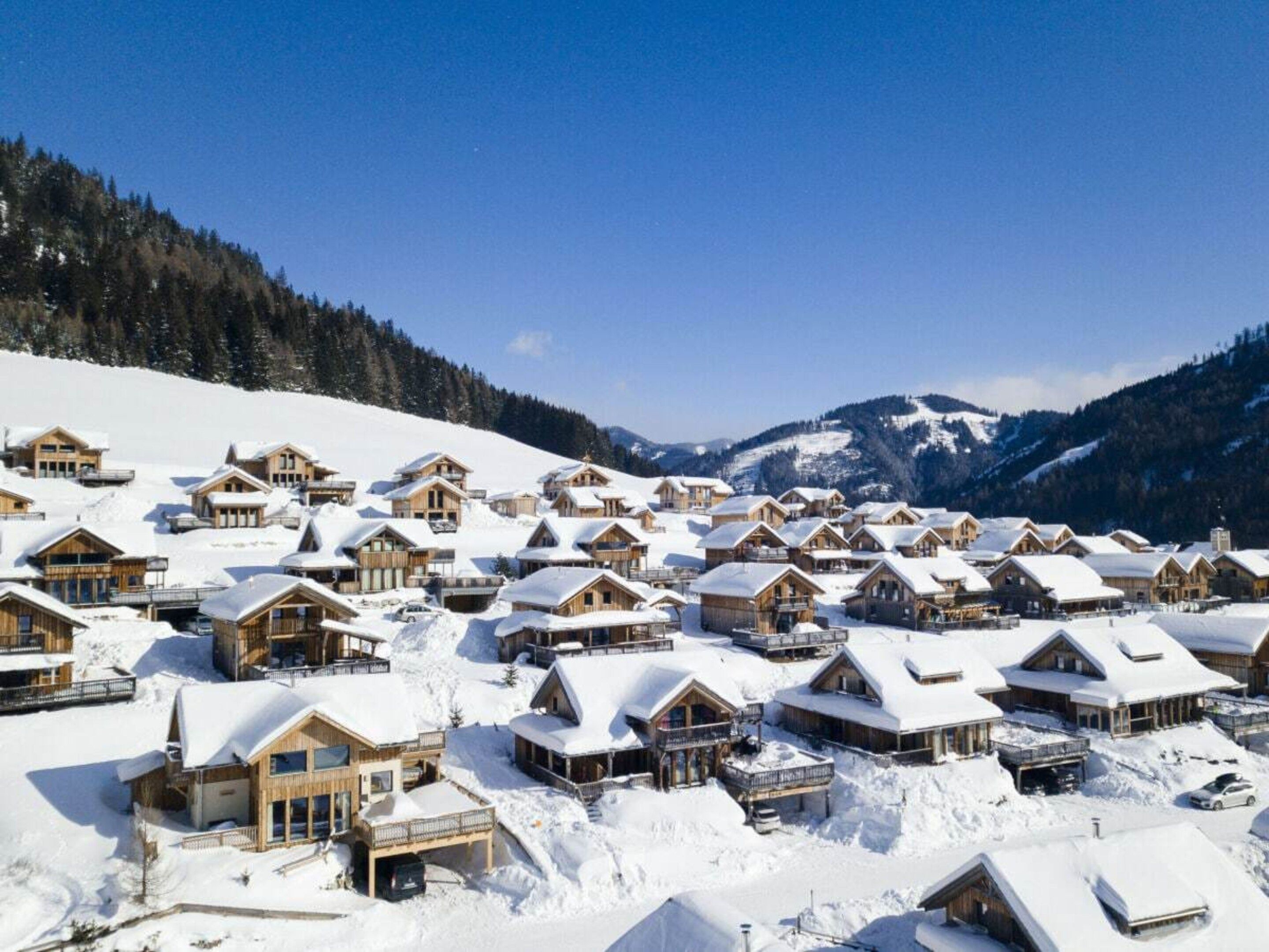 A snow-covered resort with multiple wooden cabins, a ski lift, and a mountainous backdrop.