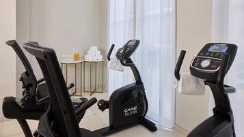A home gym with two stationary bikes, a small table with towels, and a window with curtains.