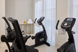 Uma academia em casa com duas bicicletas ergométricas, uma mesa pequena com toalhas e uma janela com cortinas.