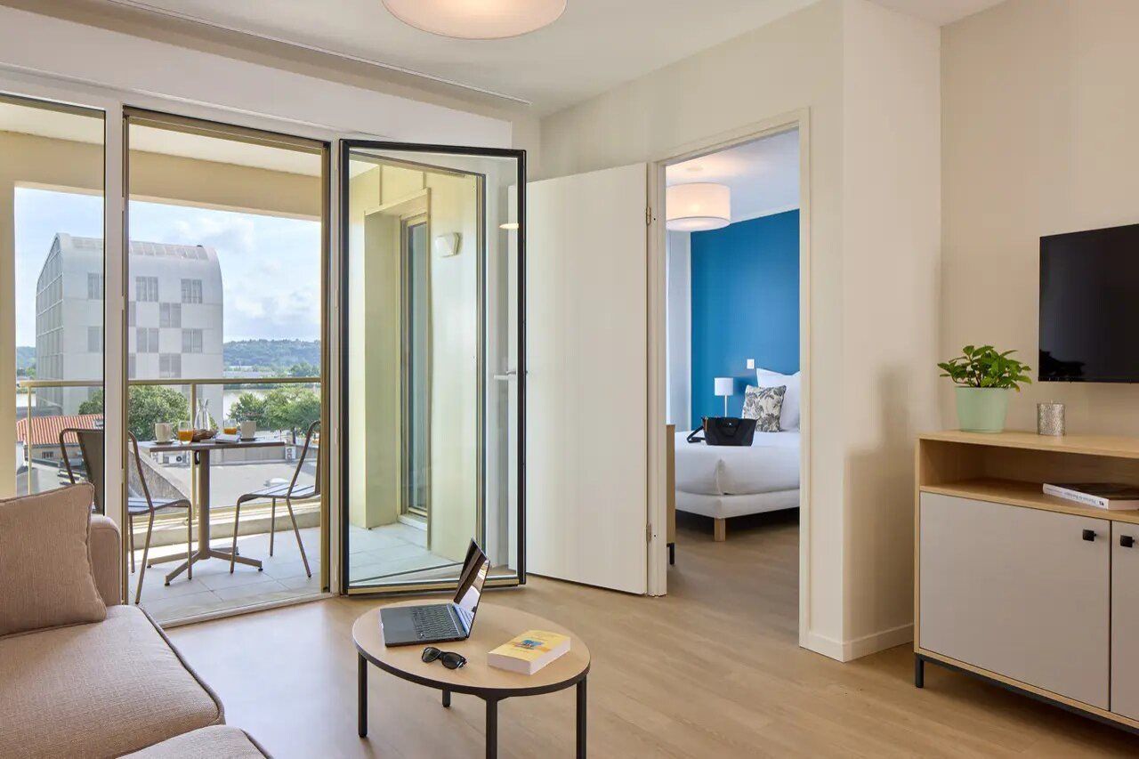 Quarto de hotel moderno com varanda, sofá, uma pequena mesa com um laptop e televisão na parede.