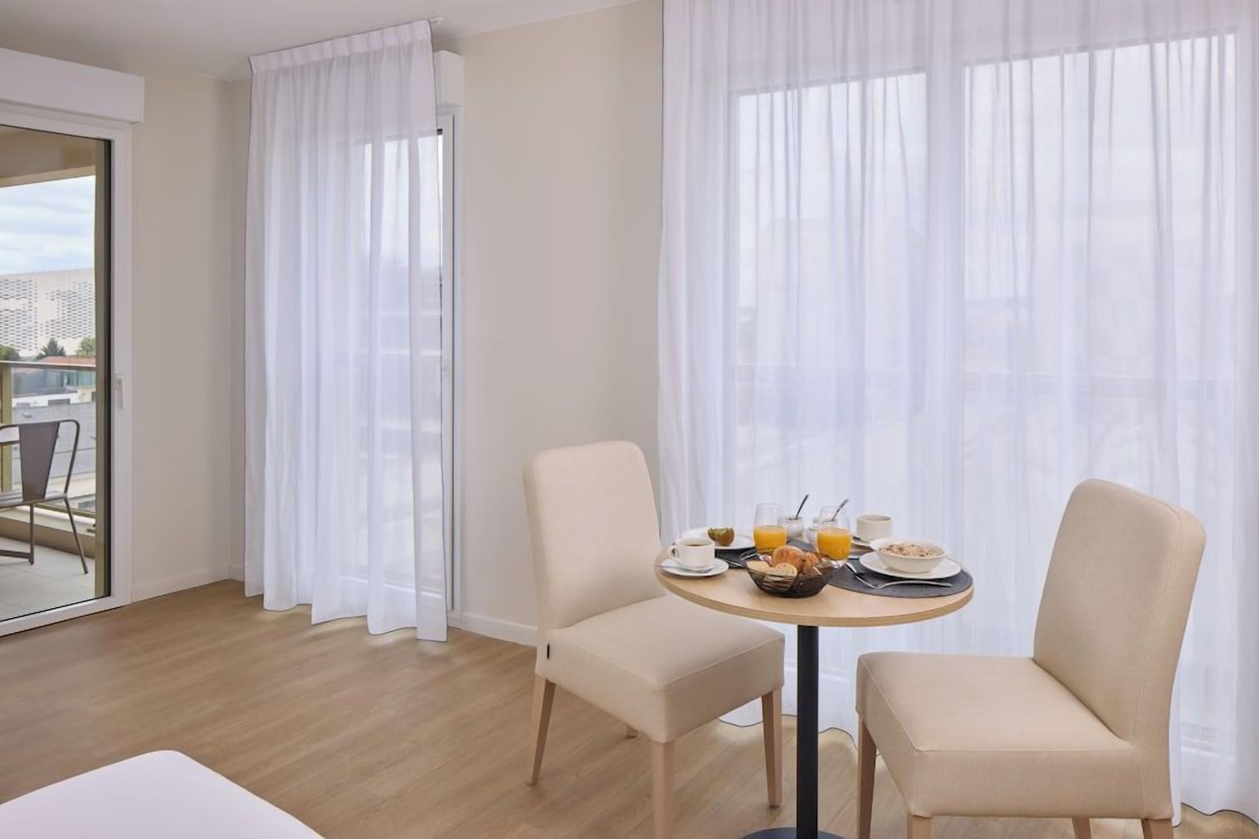 A hotel room with a dining table set for breakfast, two chairs, and a view through sheer curtains.