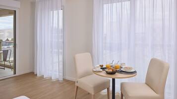 A hotel room with a dining table set for breakfast, two chairs, and a view through sheer curtains.