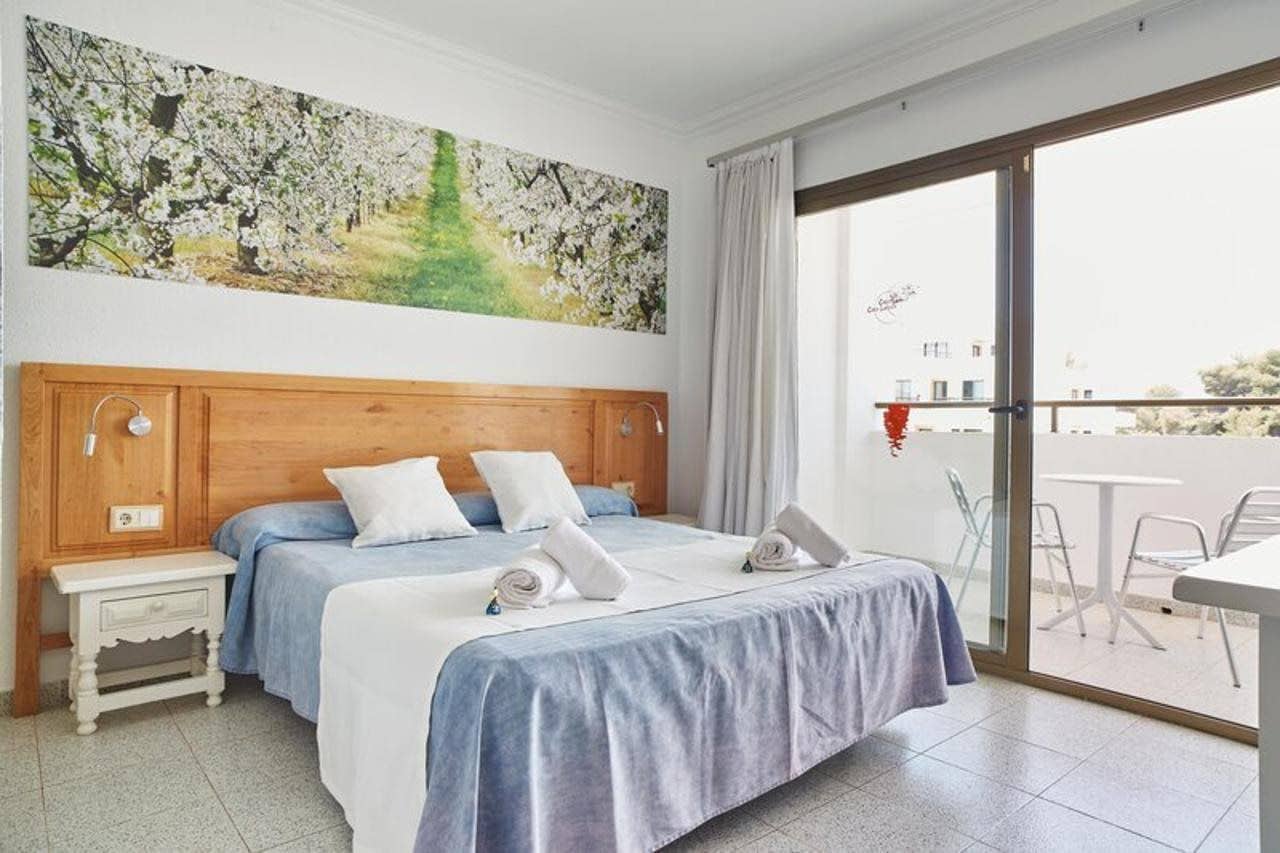 A hotel room with a large bed, a wooden headboard, a nightstand, a painting on the wall, and a balcony with outdoor seating.