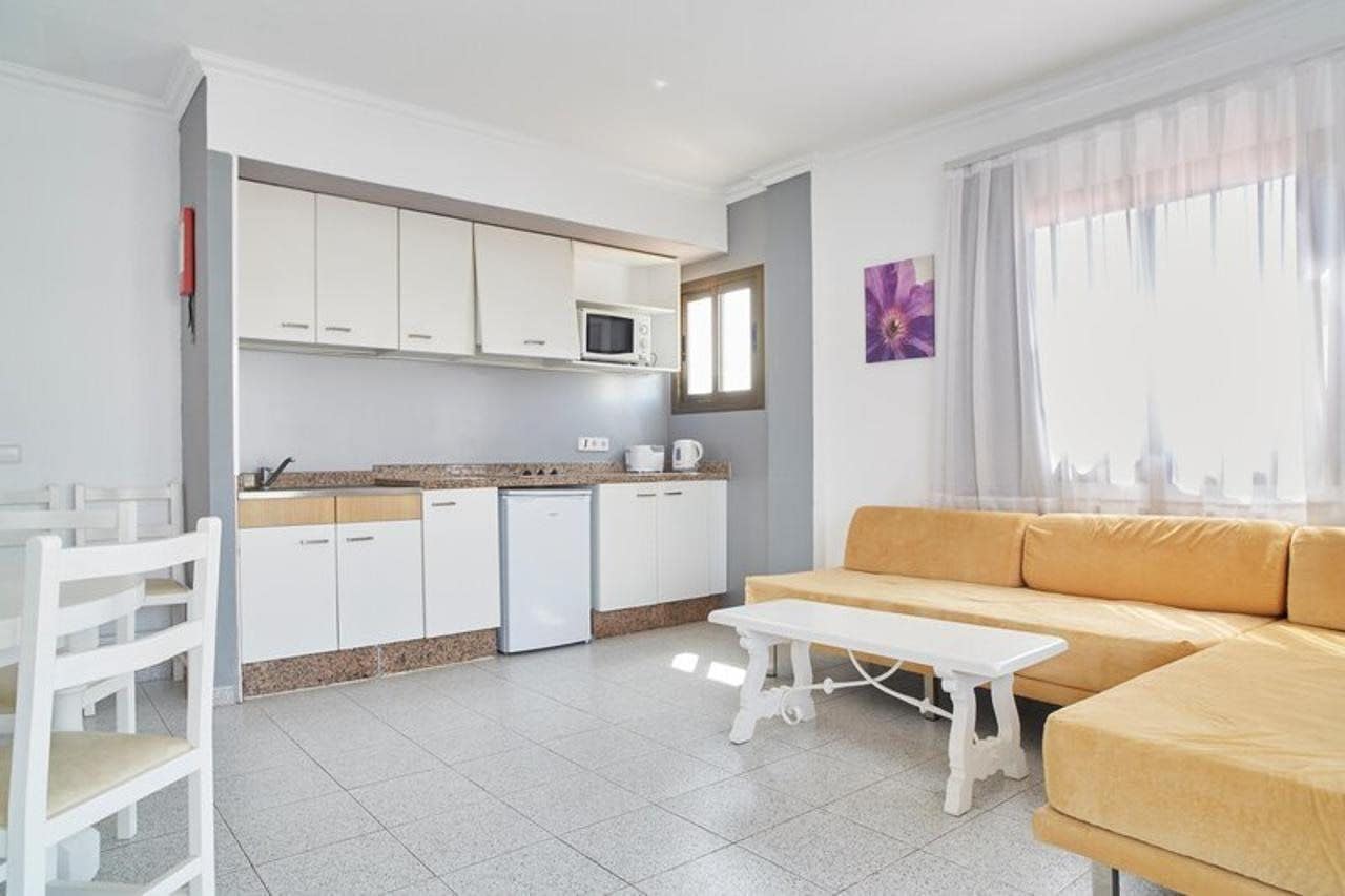 A modern kitchen with white cabinets, a microwave, and a coffee maker. There is a white dining table with chairs, a white bench, and a yellow sofa.