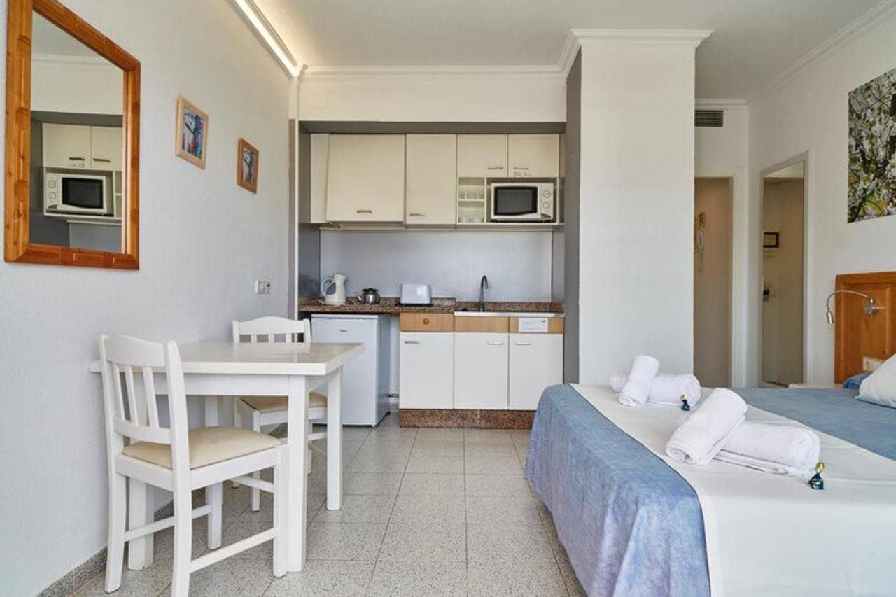 A compact hotel room with a kitchenette, a single bed with a blue bedspread, a white chair, and a small table.