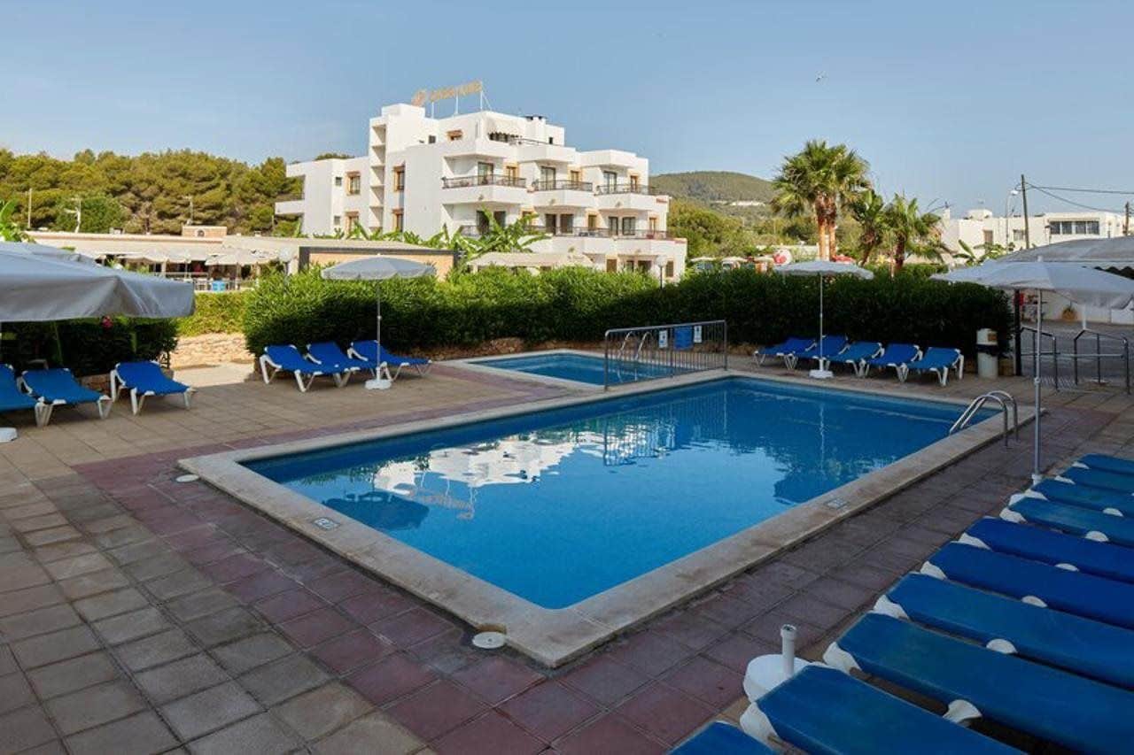 Una piscina con sillas reclinables azules y sombrillas, rodeada de un área pavimentada y un edificio al fondo.