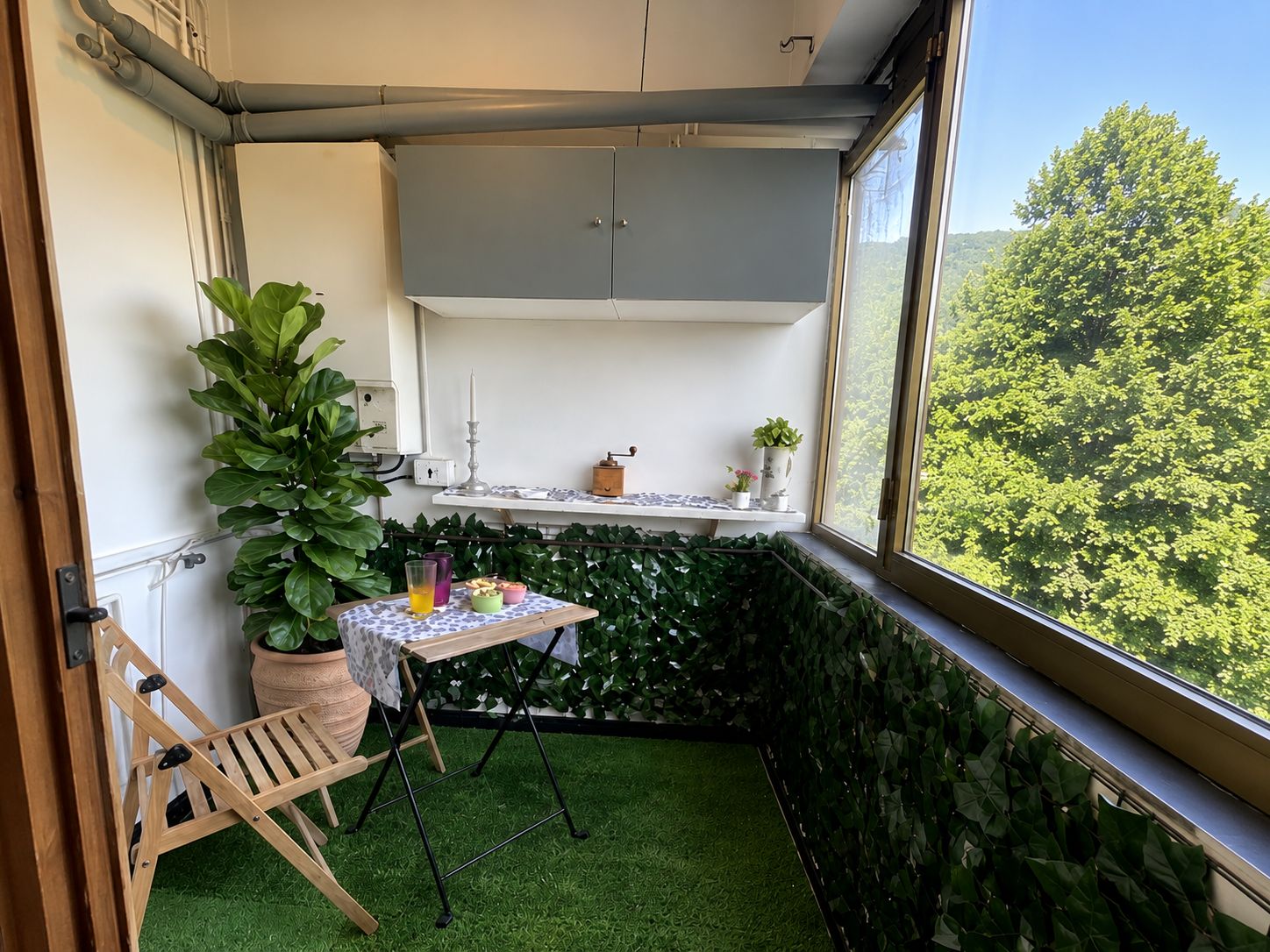 Un pequeño balcón con césped artificial, una silla de madera, una mesa con mantel blanco y plantas en macetas.