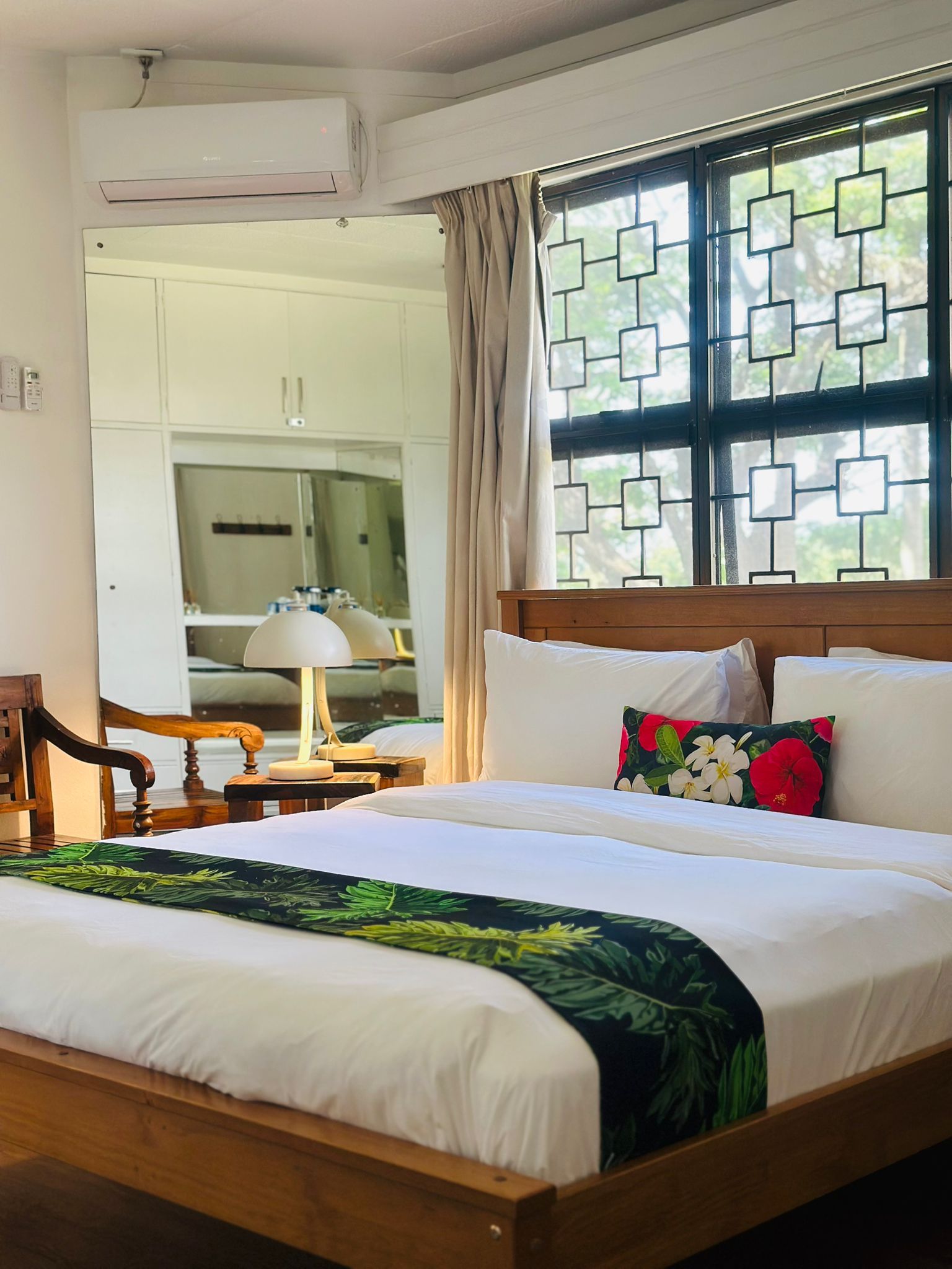 Een hotelkamer met een bed, nachtkastje, lamp, raam met geometrisch patroon en uitzicht op buiten.