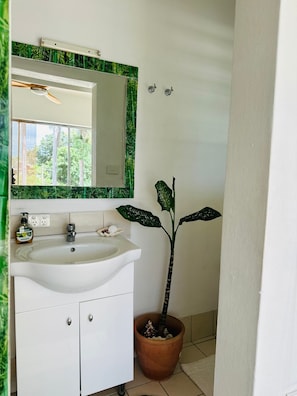 Een badkamer met een wit wastafel, een spiegel met een groene lijst, een potplant en een raam met uitzicht op groen.