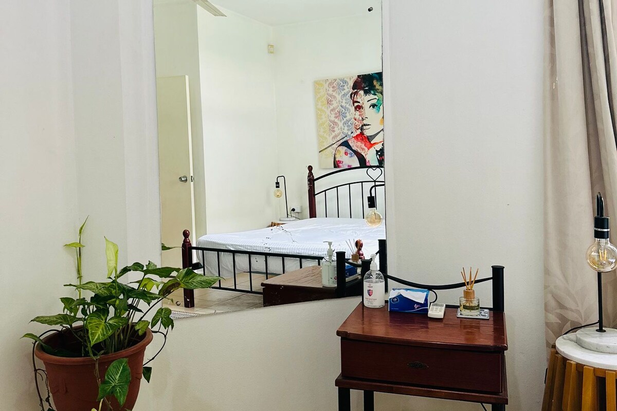 Een slaapkamer met een bed, nachtkastje, spiegel, een potplant en een kleine ronde tafel met een lamp.