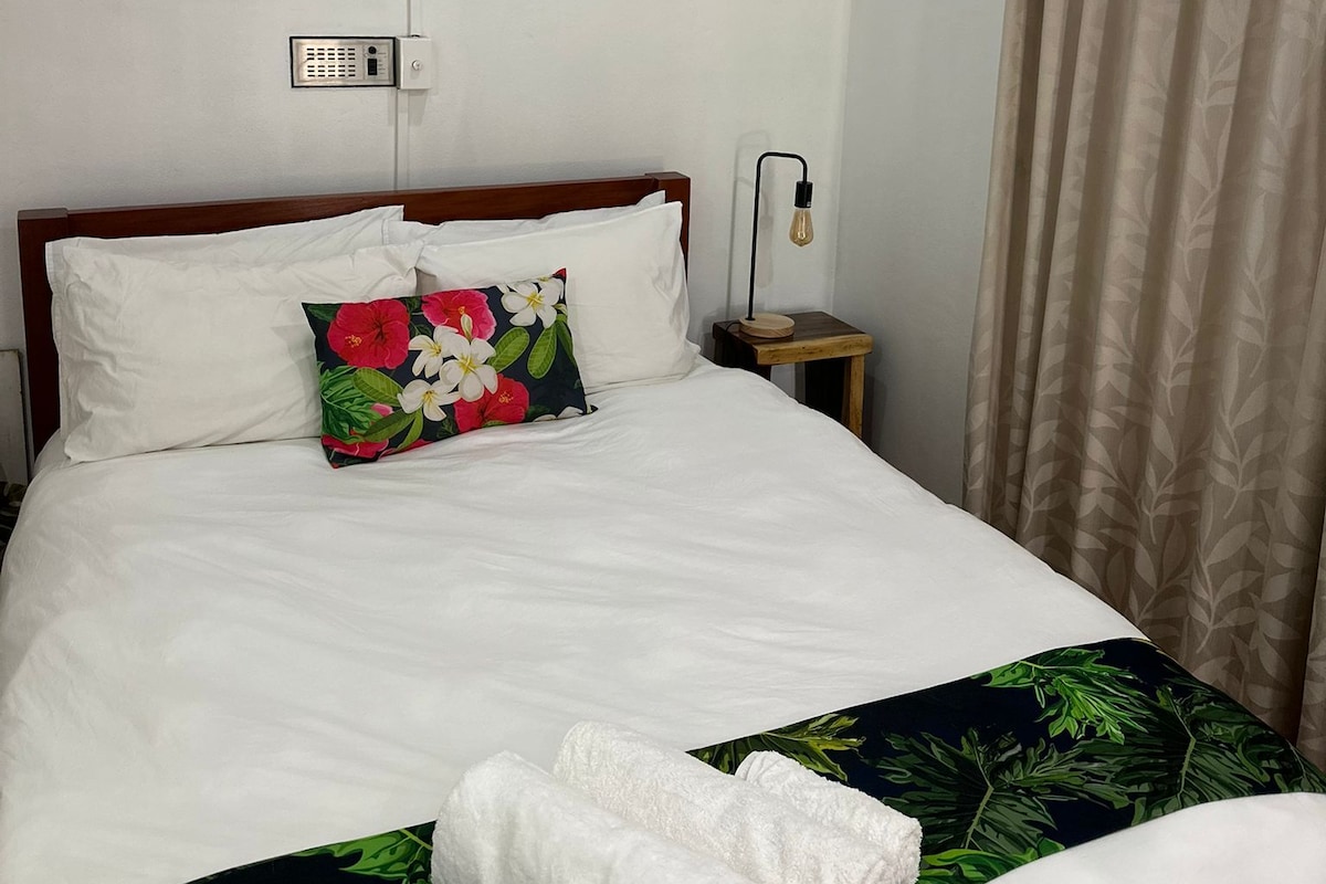 Een hotelkamer met een bed, een plafondventilator, een airconditioning, een bedlampje en een raam met gordijnen.