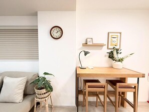 Ein Zimmer mit einem Holztisch, zwei Stühlen, einem kleinen Topfpflanzen und einer Wanduhr sowie einem gerahmten Bild.