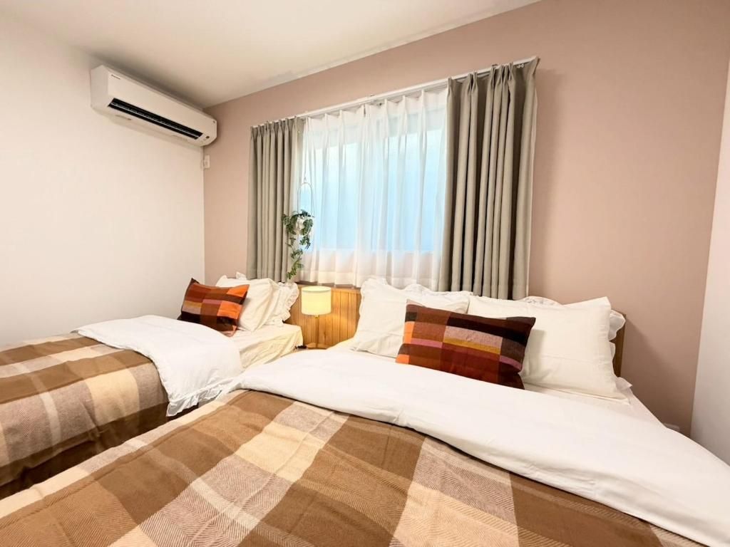 A hotel room with two beds, a bedside table with a lamp, a window with curtains, and an air conditioning unit.