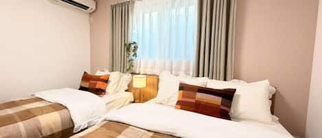 A hotel room with two beds, a bedside table with a lamp, a window with curtains, and an air conditioning unit.