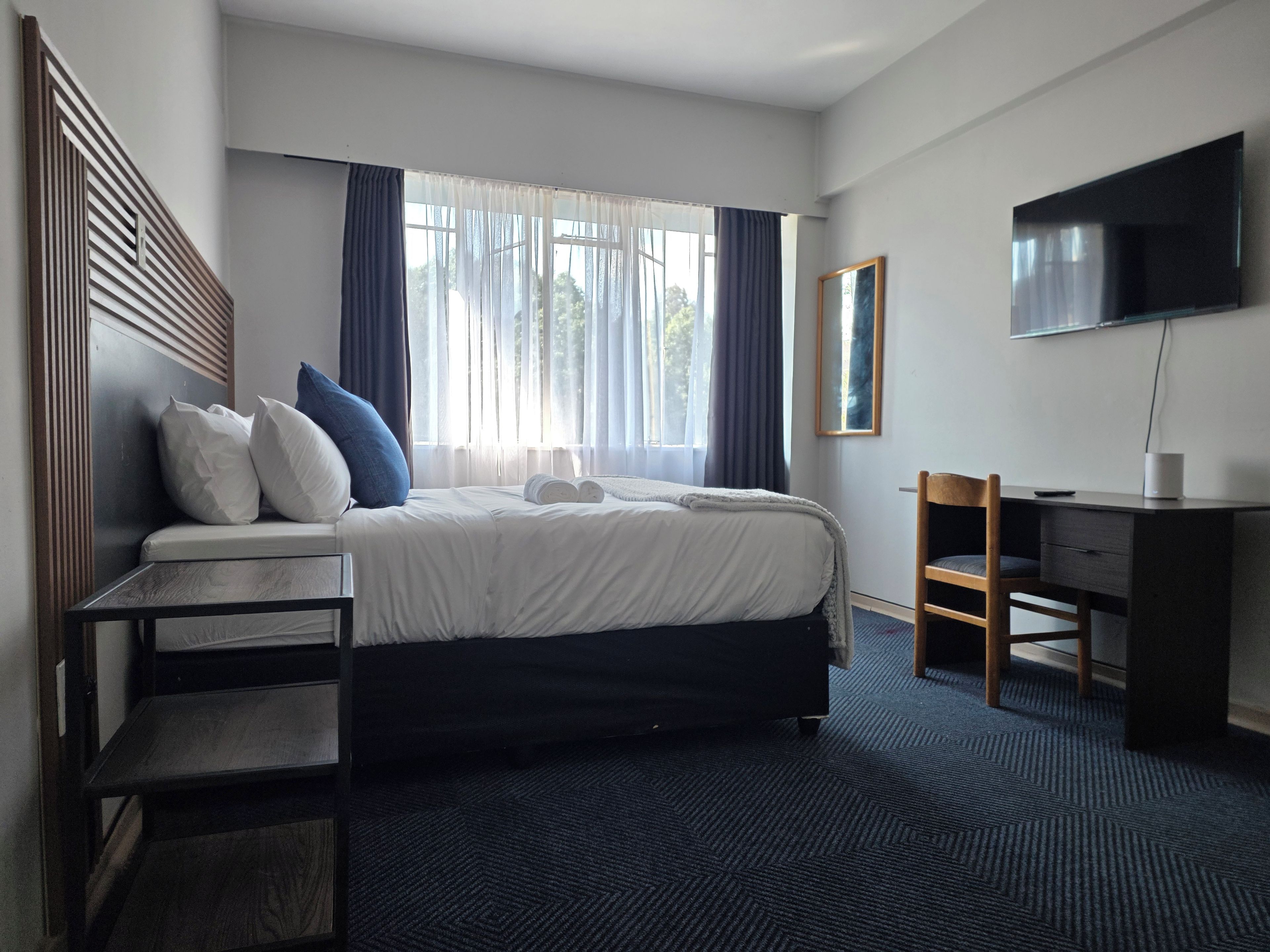 Habitación de hotel con cama, escritorio, silla, televisor y ventana con cortinas.