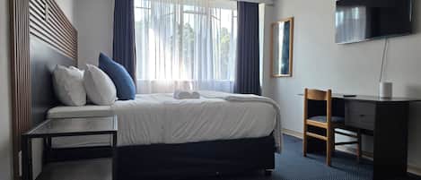 Quarto de hotel com cama, escrivaninha, cadeira, televisão e janela com cortinas.