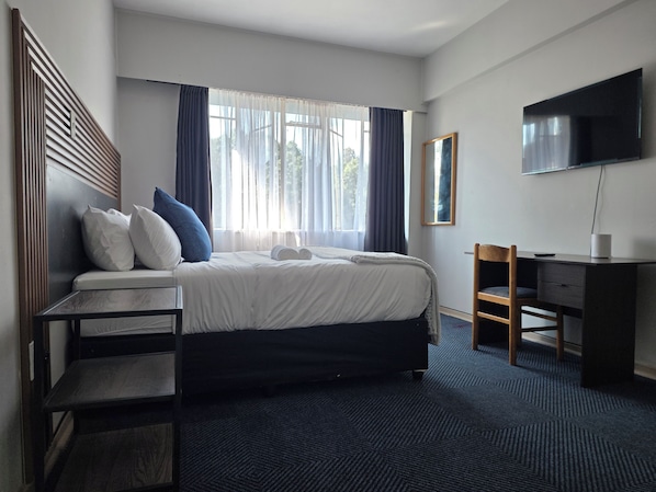 Habitación de hotel con cama, escritorio, silla, televisor y ventana con cortinas.