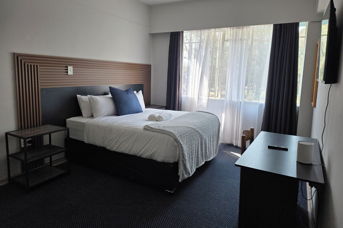 A hotel room with a bed, bedside table, and a desk with a chair.