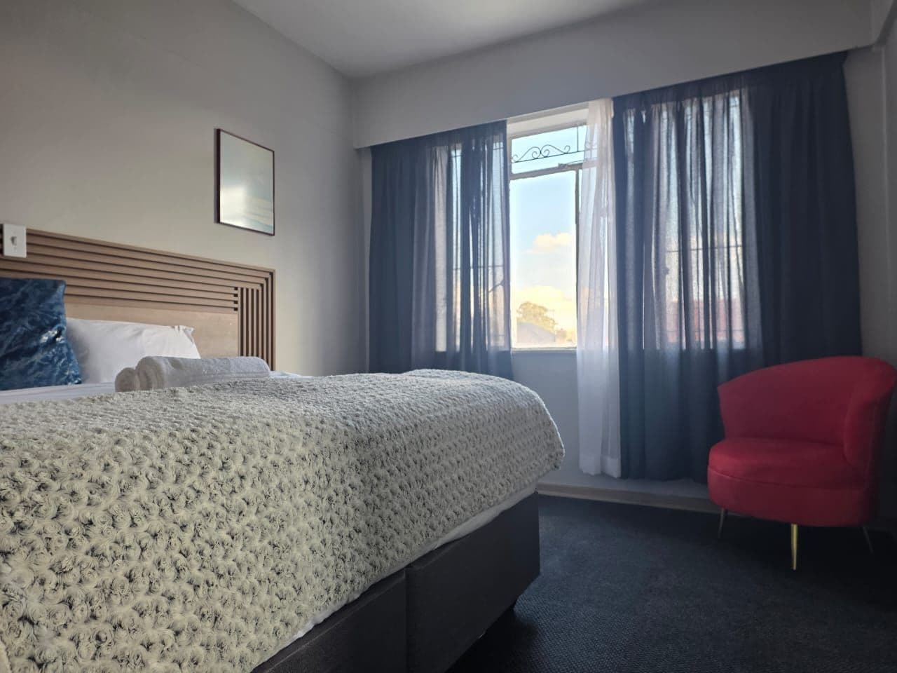 A hotel room with a bed, a red chair, a window with curtains, and a framed picture on the wall.