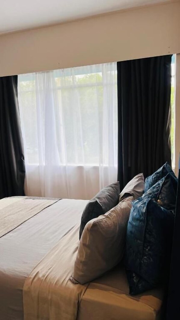 A bed with a beige cover and two pillows, a window with sheer white curtains, and a view of greenery outside.