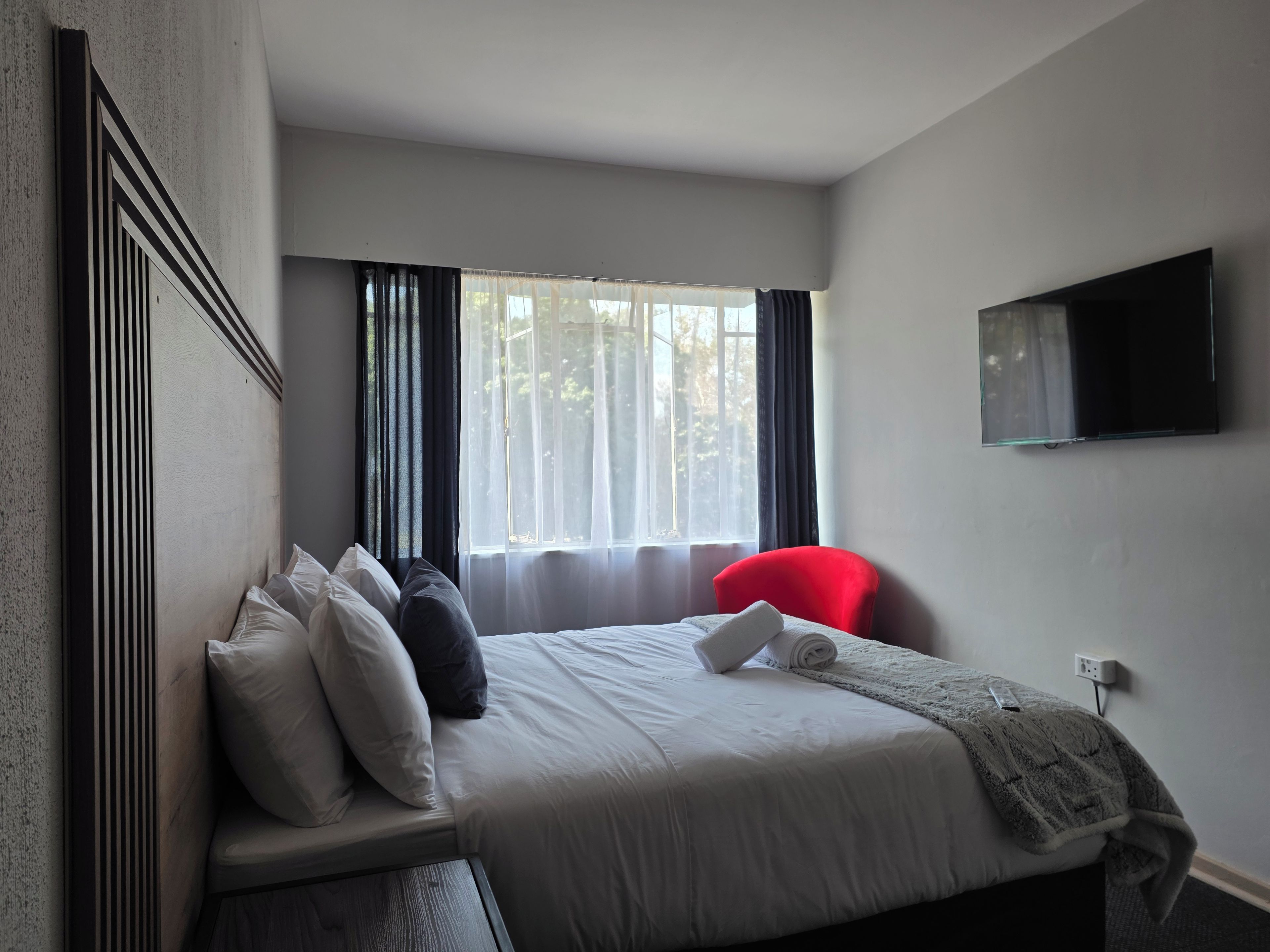 Una habitación de hotel con una cama, una silla roja, un televisor fijado a la pared y una ventana con cortinas.