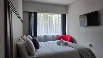 Una habitación de hotel con una cama, una silla roja, un televisor fijado a la pared y una ventana con cortinas.