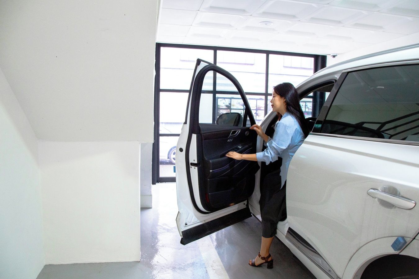A person opening a car door in a well-lit indoor space with a large window in the background.