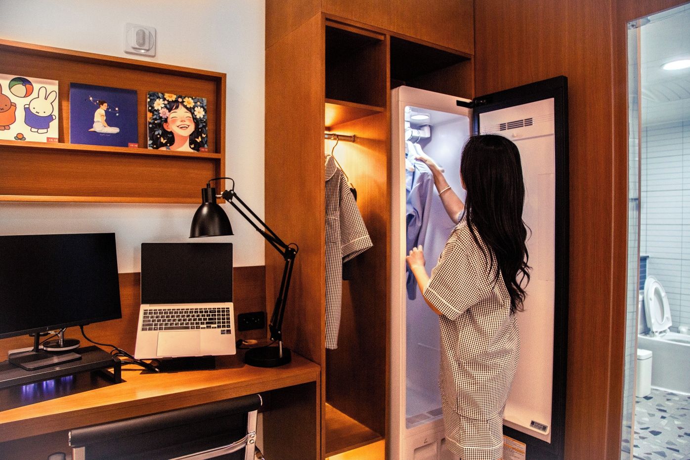 A hotel room with a desk, computer, lamp, and a wardrobe with clothes.