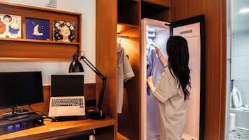 A hotel room with a desk, computer, lamp, and a wardrobe with clothes.