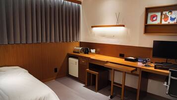 A hotel room with a bed, a desk with a computer, a small refrigerator, and a bedside table.