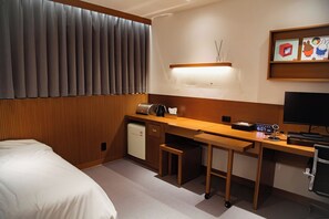 Una habitación de hotel con una cama, un escritorio con computadora, un refrigerador pequeño y una mesita de noche.