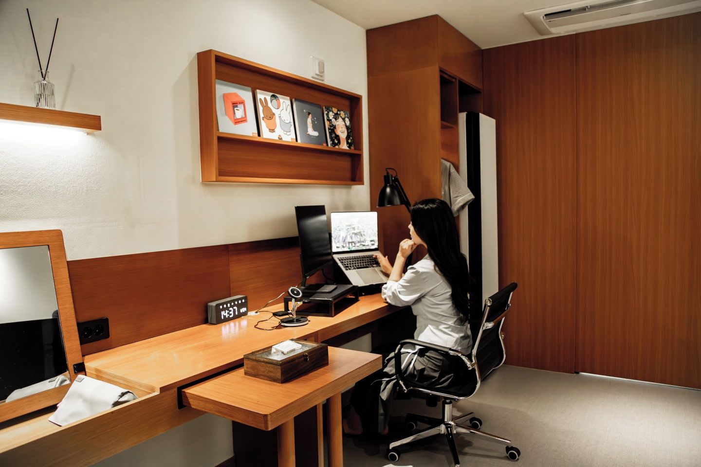 A modern office space with a wooden desk, a laptop, a desktop computer, and a shelf with framed artwork.