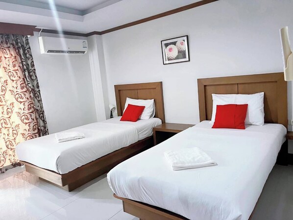 A hotel room with two single beds, each with a wooden headboard, white bedding, and a red pillow. There is a framed picture on the wall above the beds, and an air conditioning unit mounted on the wall. - Al Ameen Hotel (Krabi)