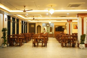 A hotel dining area with multiple wooden tables and chairs, ceiling fans, and a chandelier. - Sunrise Resorts (jaisalmer)