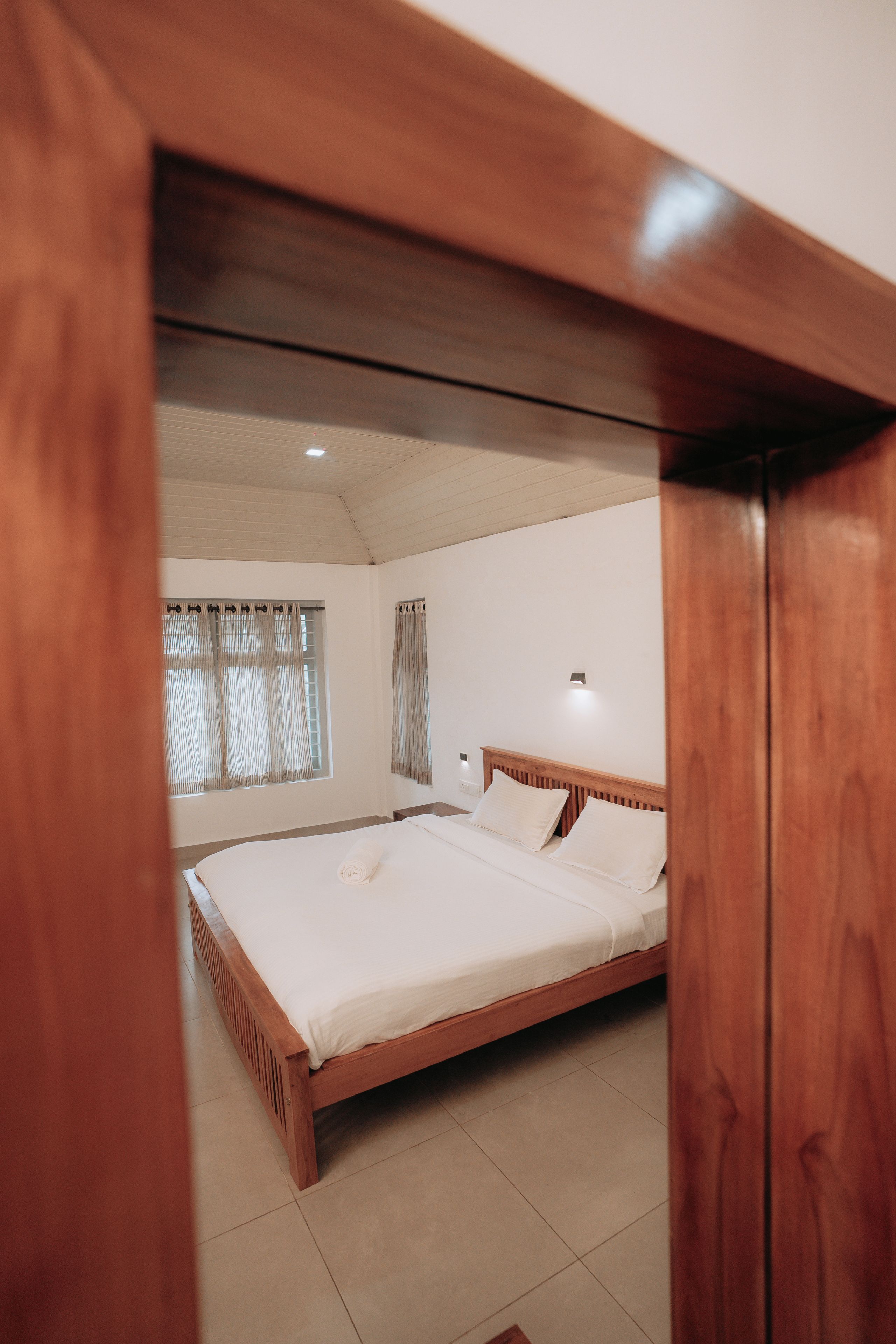 Ein Schlafzimmer mit einem hölzernen Bett, weißen Leinentüchern, einem Nachttisch, einer Wandlampe und einem Fenster mit Vorhängen.