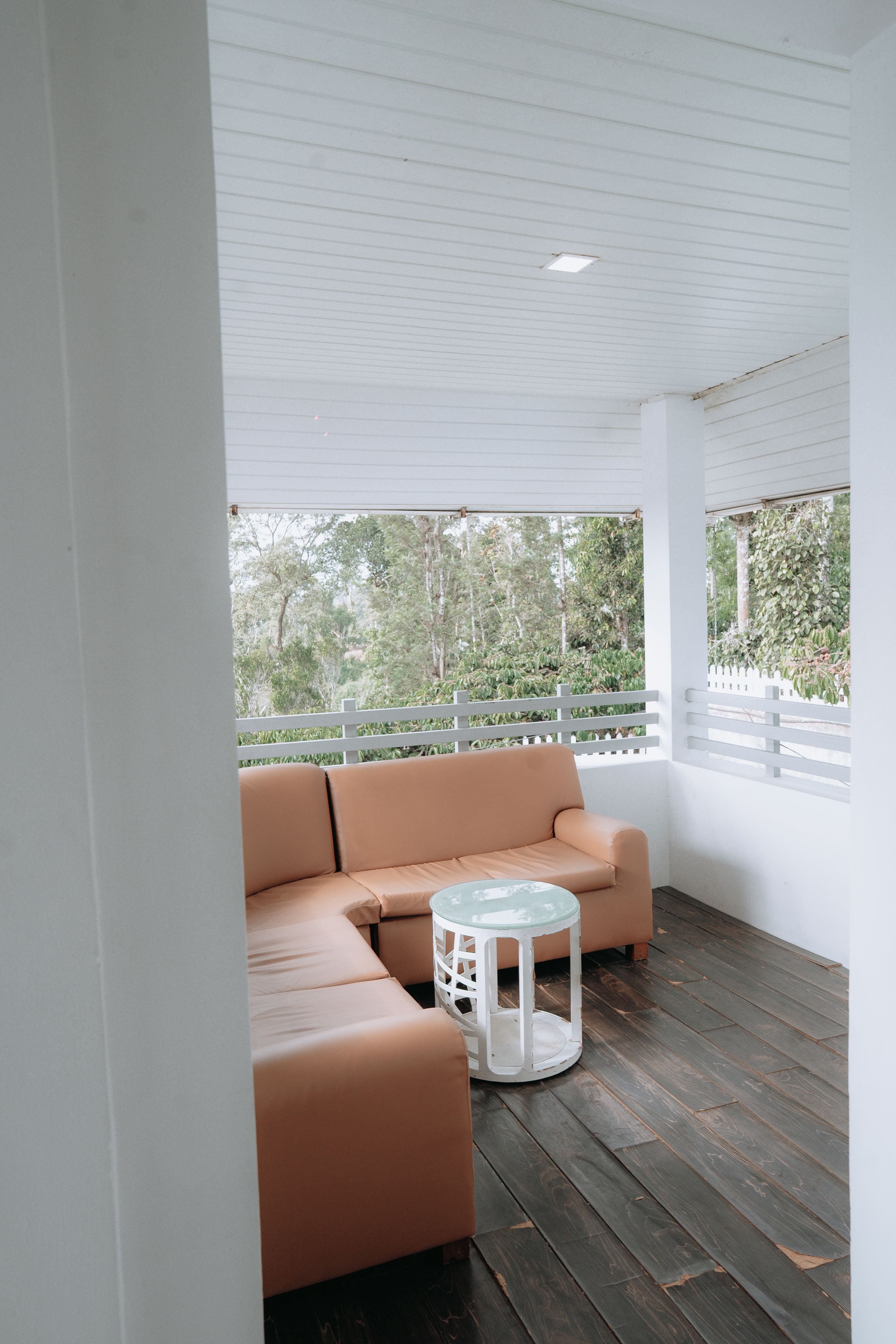 Un espacio moderno al aire libre con sofás y una pequeña mesa redonda, suelo de madera y vistas a zonas verdes a través de una barandilla blanca.