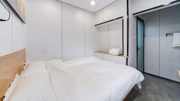 A modern hotel room with a bed, a wooden headboard, a wall-mounted shelf with a towel, and a door leading to a bathroom.