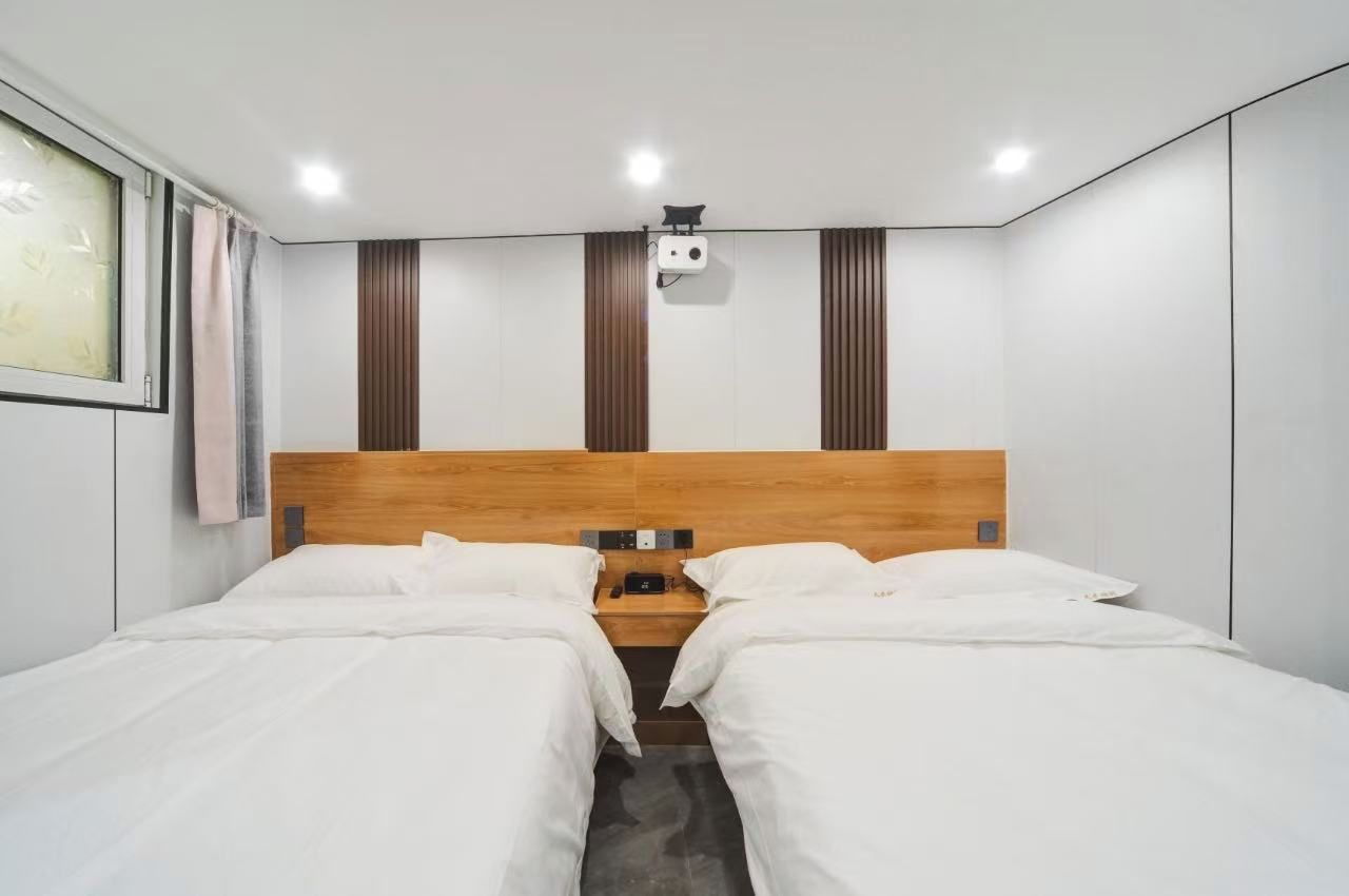 Quarto de hotel moderno com cabeceira de madeira, duas camas, uma mesa de cabeceira e projetor fixado no teto.
