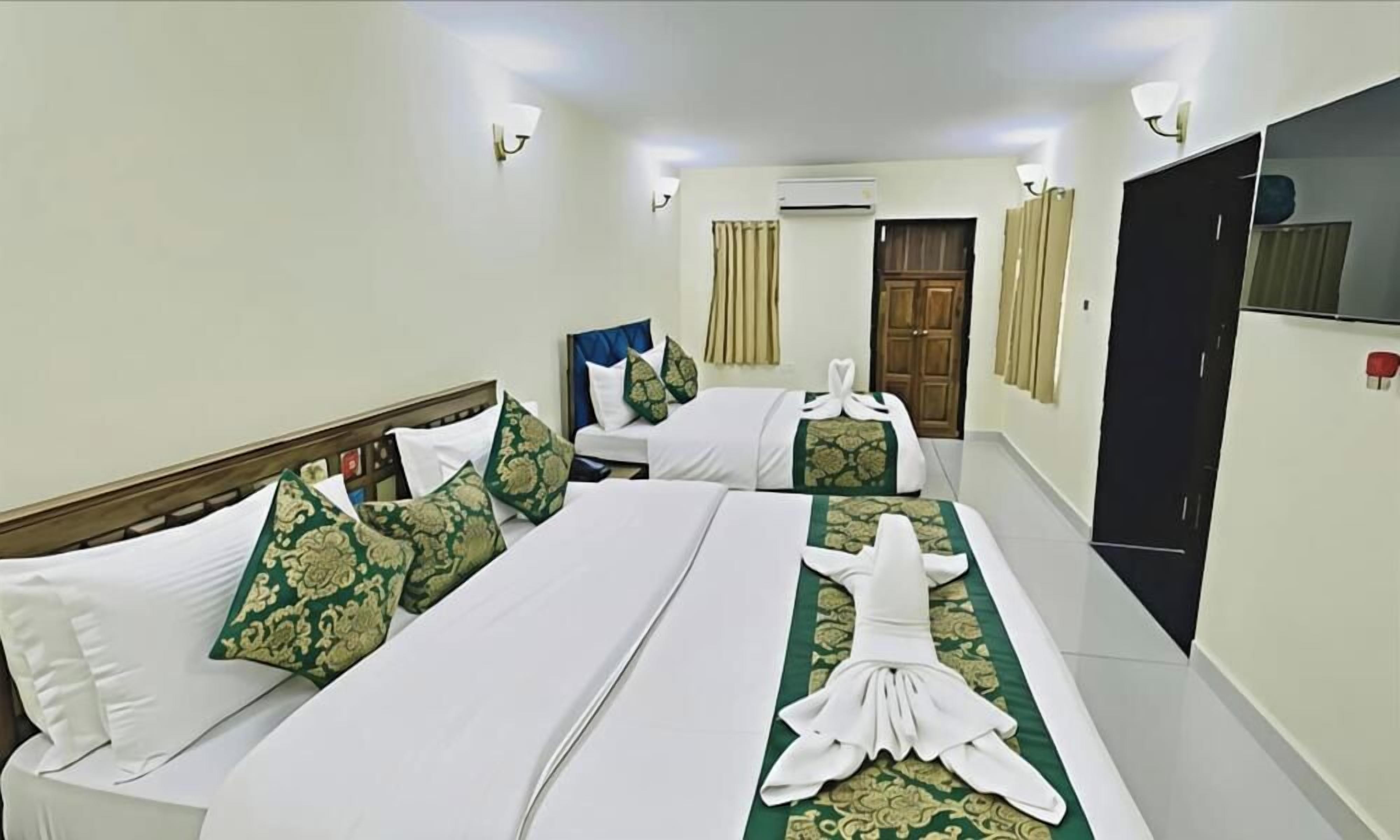 A hotel room with two beds, each with white linens and green patterned pillows. There is a flat-screen TV on the wall and a wardrobe.