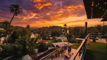 Terraza en la azotea con vista a un atardecer sobre un paisaje urbano, que incluye palmeras y edificios.