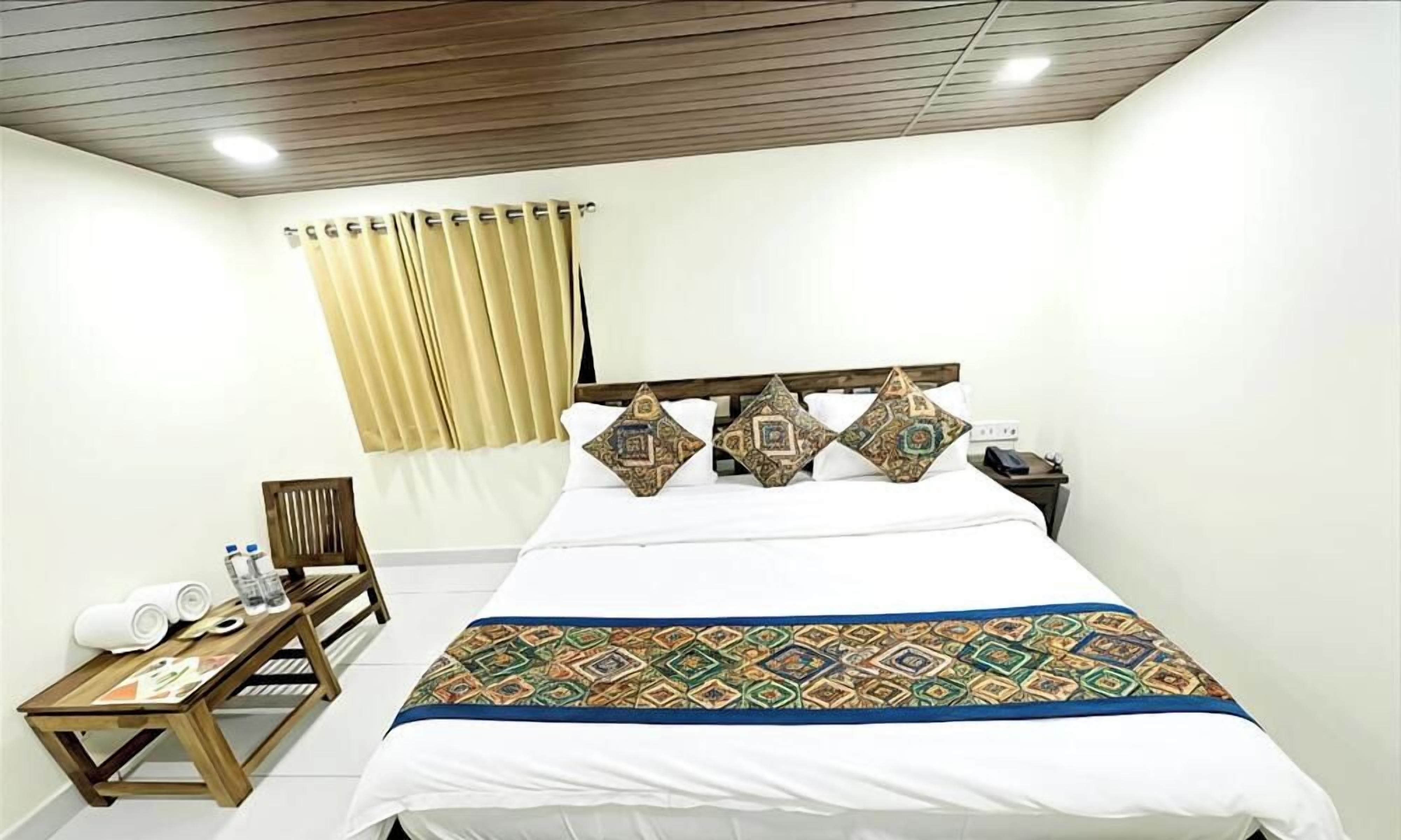 A hotel room with a large bed, a wooden nightstand, a chair, a small table with towels and a water bottle, and a window with curtains.