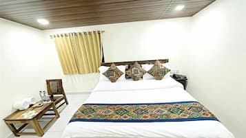A hotel room with a large bed, a wooden nightstand, a chair, a small table with towels and a water bottle, and a window with curtains.
