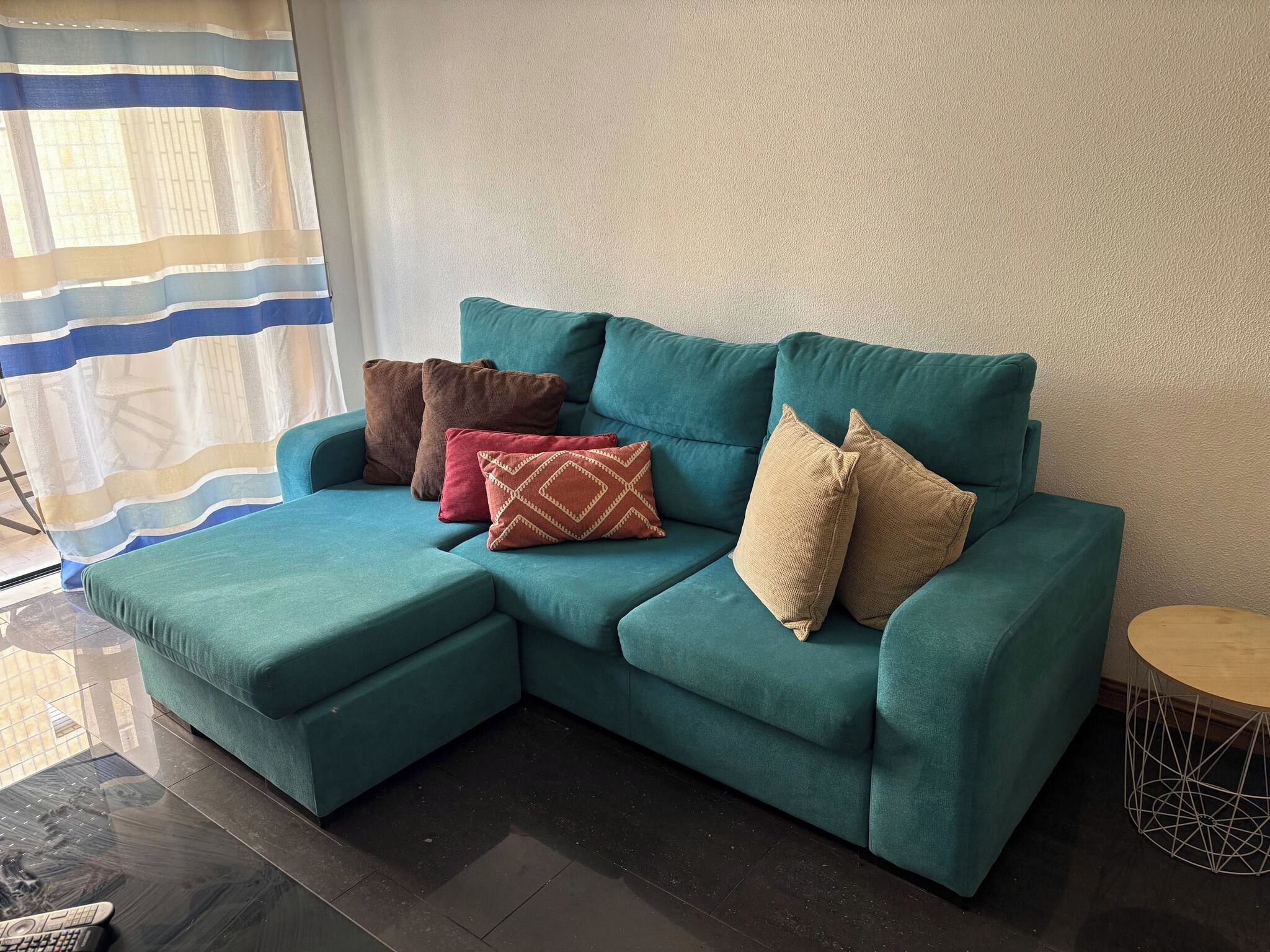 Un canapé d’angle de couleur turquoise, accompagné d’un pouf assorti, de trois coussins à motifs, d’une petite table ronde et d’un rideau à rayures.