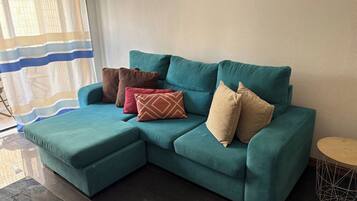 Un canapé d’angle de couleur turquoise, accompagné d’un pouf assorti, de trois coussins à motifs, d’une petite table ronde et d’un rideau à rayures.