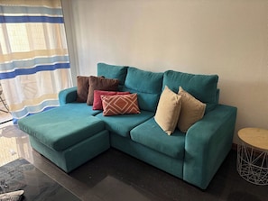 A teal sectional sofa with a matching ottoman, three patterned cushions, a small round table, and a striped curtain.