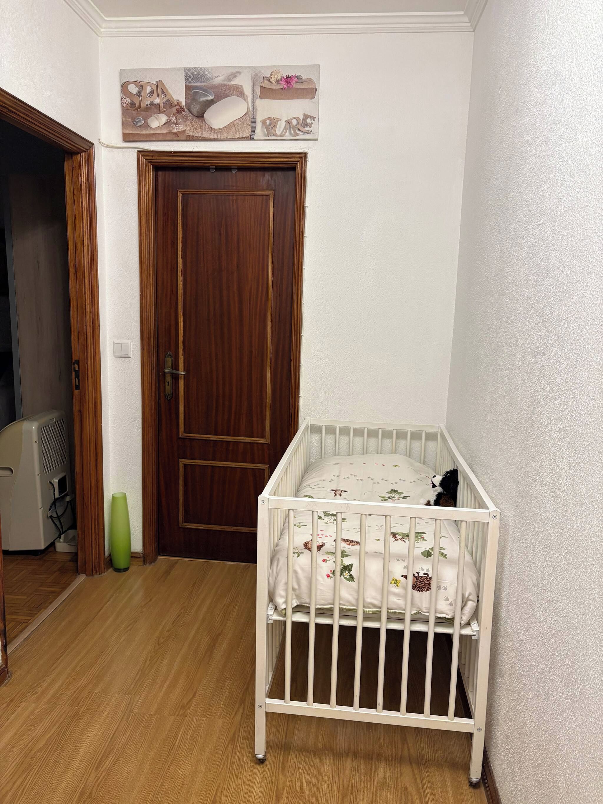 Una habitació amb una porta de fusta, una bressol blanca amb roba de llit floral, i un vas verd.