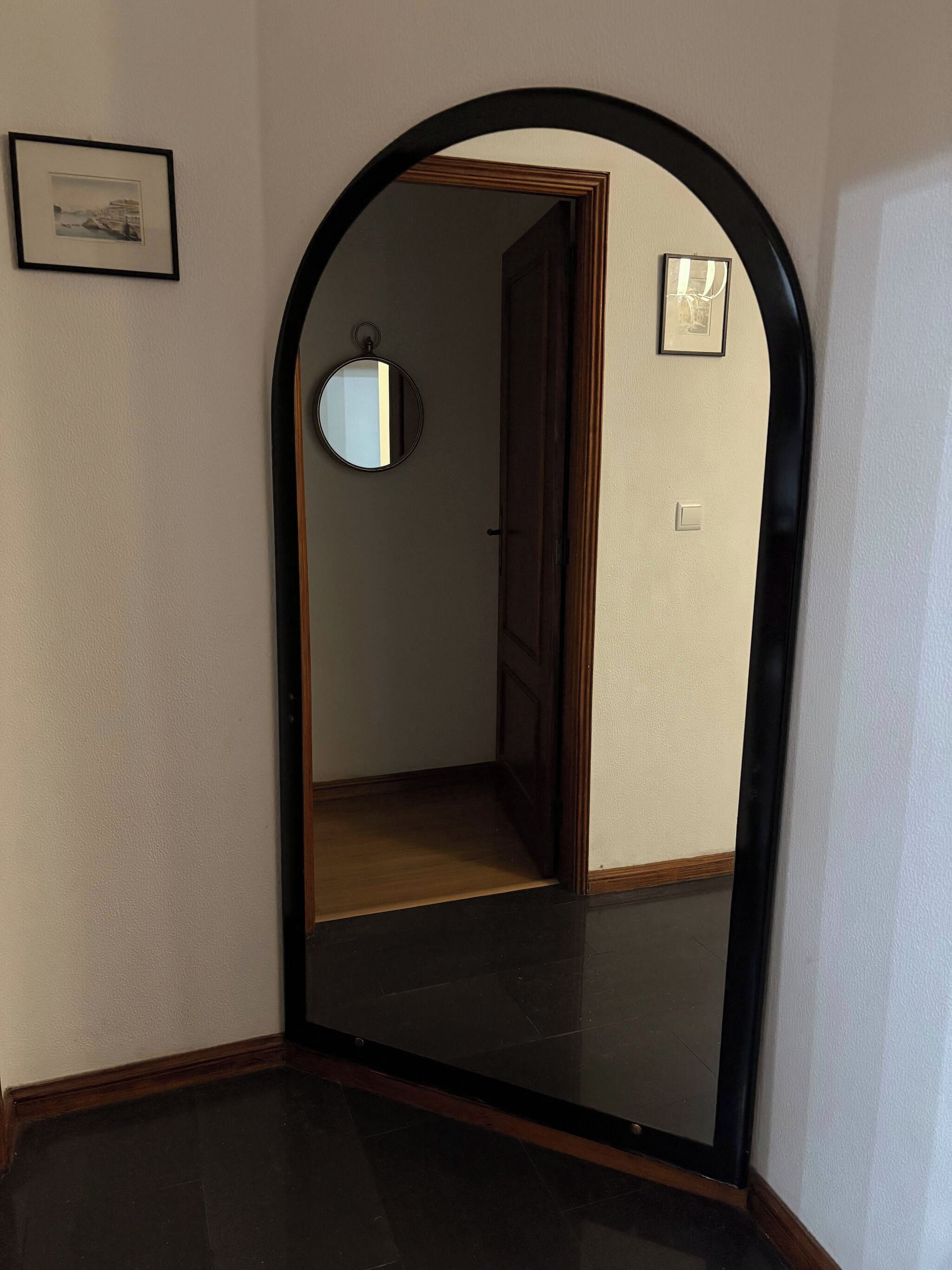 Un mirall amb arc que reflecteix un passadís amb una porta, un quadre amb marc a la paret i un interruptor de llum.