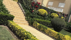 Un immeuble résidentiel à plusieurs étages, doté d’un escalier menant à l’entrée, et entouré de jardins et de haies bien entretenus.